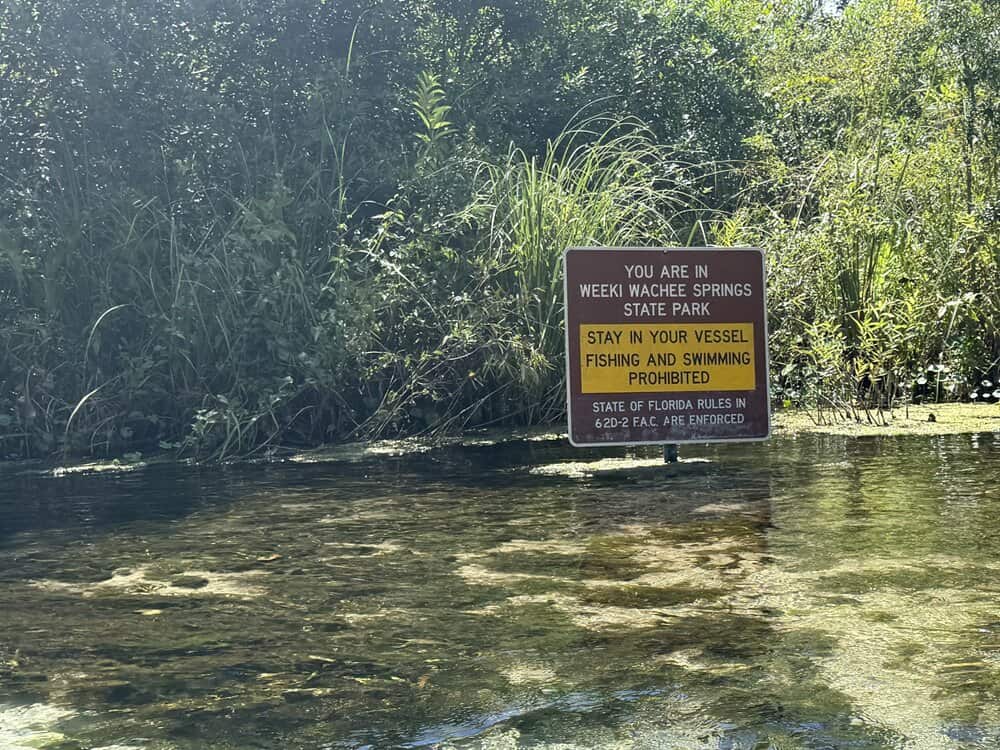 Flooded area at Weeki Wachee Springs State Park with warning sign about vessel restrictions and swimming prohibition.