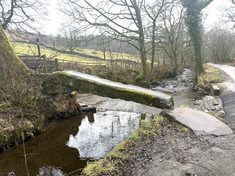 Scenic view of Wycoller village stream with a small footbridge and rural pathway, surrounded by leafless trees and countryside in England.